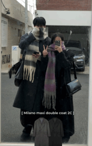 Корейская пара в длинных пальто и тёплых шарфах позирует у зеркала на улице зимой